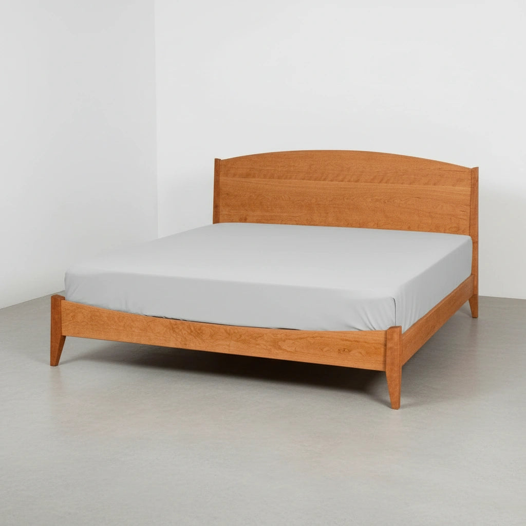 The Shaker Platform Bed - Solid Wood Bed Frame by T.Y. Fine Furniture features a curved headboard, tapered legs, a light brown finish, and a light gray fitted sheet, displayed against a white wall with a gray floor.