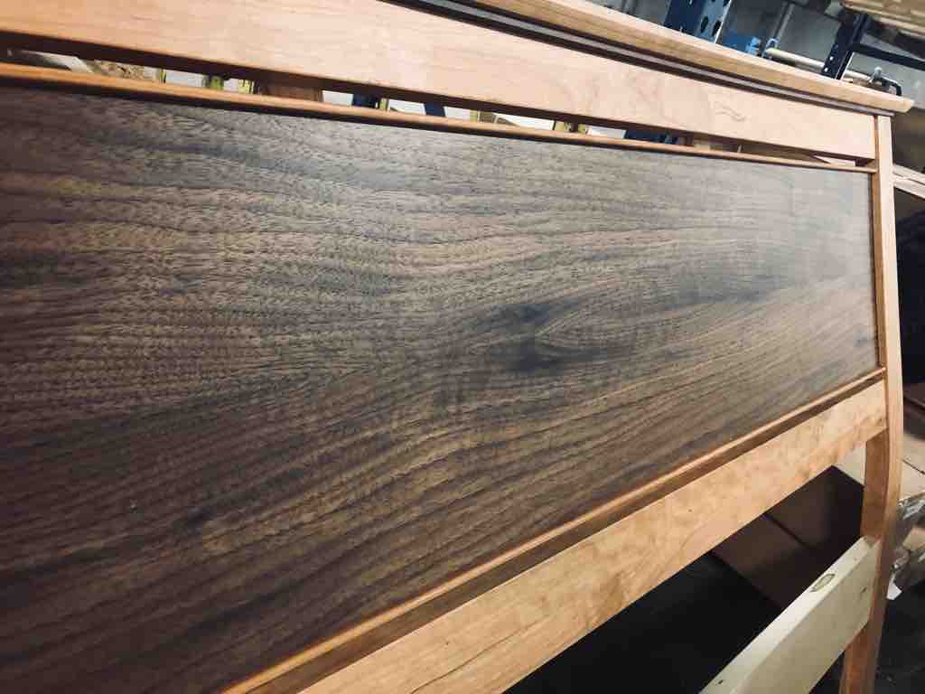 Close-up of the Enso Platform Bed- Cherry by TY Fine Furniture, showing a dark walnut panel headboard framed in cherry wood, set in a workshop with shelves and tools visible in the background.