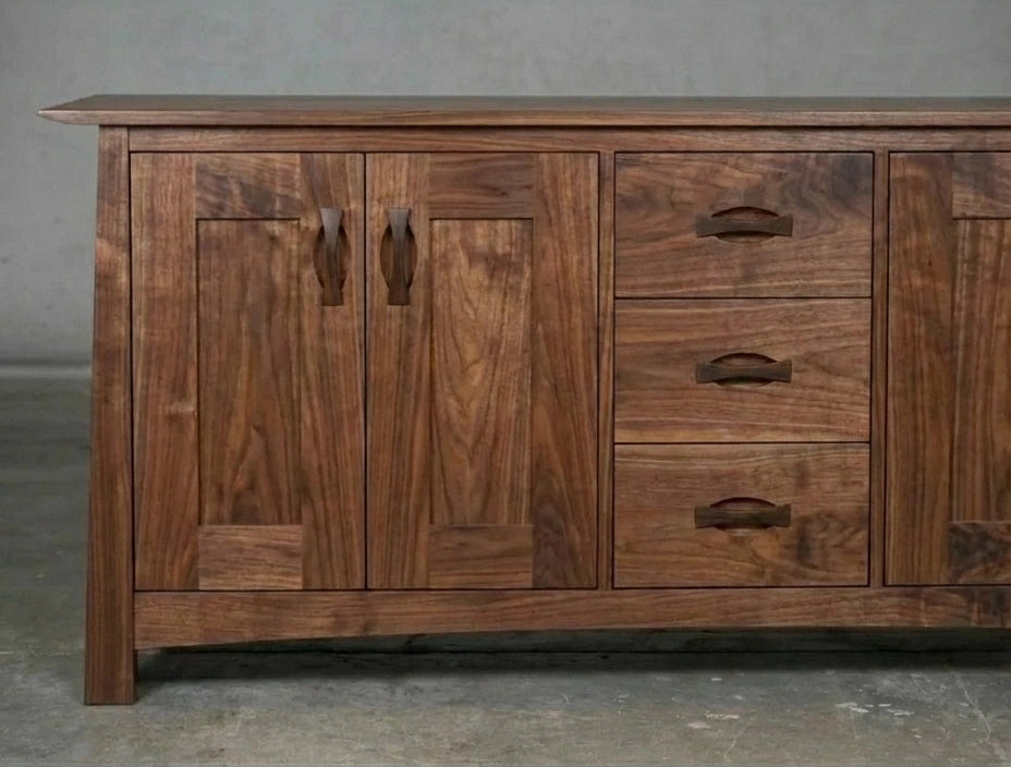 The Enso Solid Wood Credenza Buffet by TY Fine Furniture is handmade from walnut and features two paneled doors, three drawers with curved handles, and a natural finish—perfect for stylish, sustainable dining room storage.