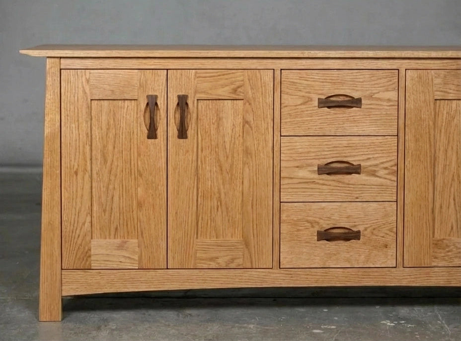 The Enso Solid Wood Credenza Buffet by TY Fine Furniture is handmade from white oak with two cabinet doors and four drawers. Featuring dark wood handles and a natural finish, it offers stylish dining room storage against a gray wall on a concrete floor.