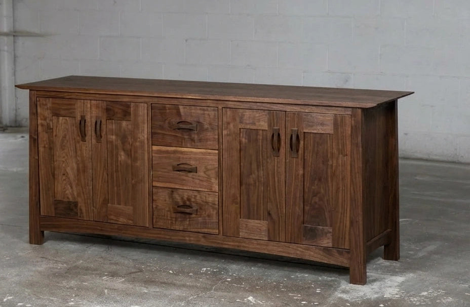 The Enso Solid Wood Credenza Buffet by TY Fine Furniture, handmade from walnut, features two side cabinets and three center drawers for dining room storage, and stands on a concrete floor against a plain gray wall.
