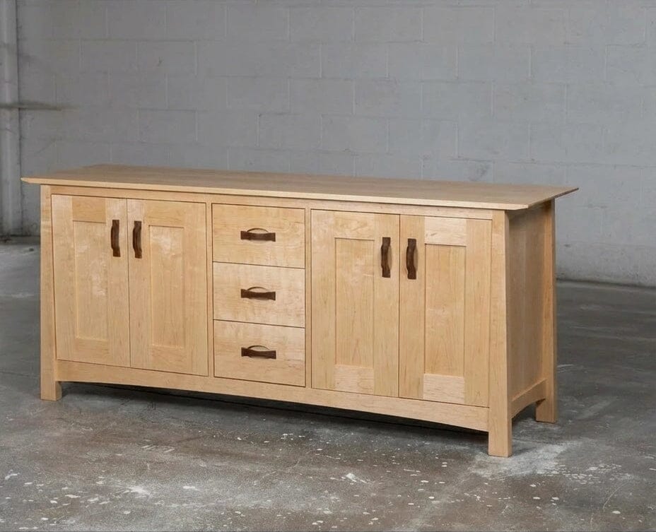 The Enso Credenza & Buffet in Maple by TY Fine Furniture features four central drawers and two double-door cabinets, providing stylish dining room storage against a plain concrete wall and floor.