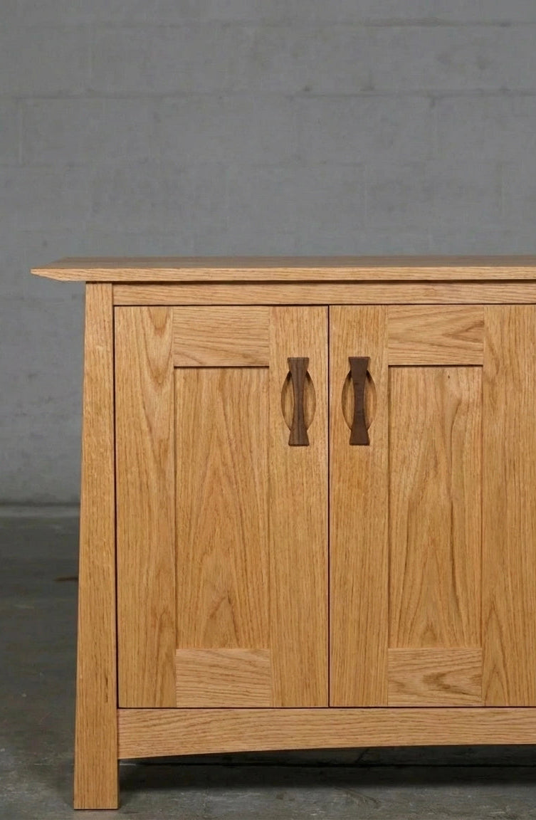 The left side of the Enso Solid Wood Credenza Buffet by TY Fine Furniture, handmade from white oak, stands against a gray wall and concrete floor, showcasing its paneled doors and curved handles for stylish dining room storage.