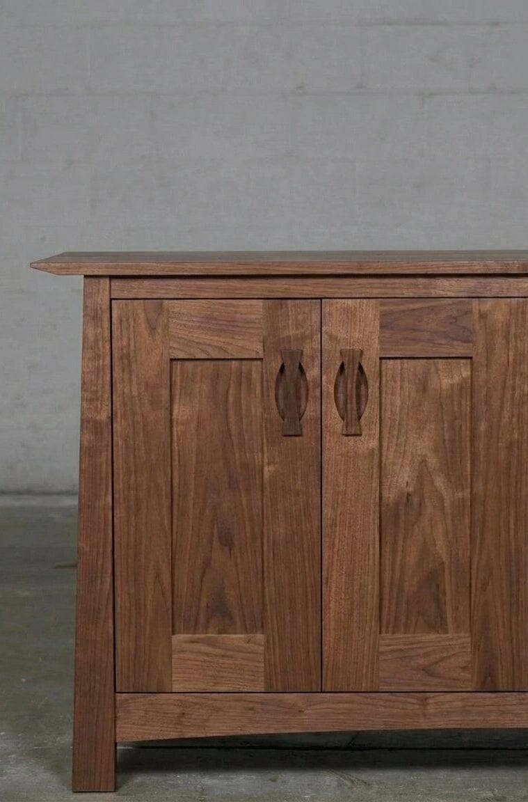 The Enso Credenza & Buffet in Walnut by TY Fine Furniture features solid wood with two paneled doors and carved handles, providing sustainable and elegant dining room storage against a plain, light-colored brick wall.