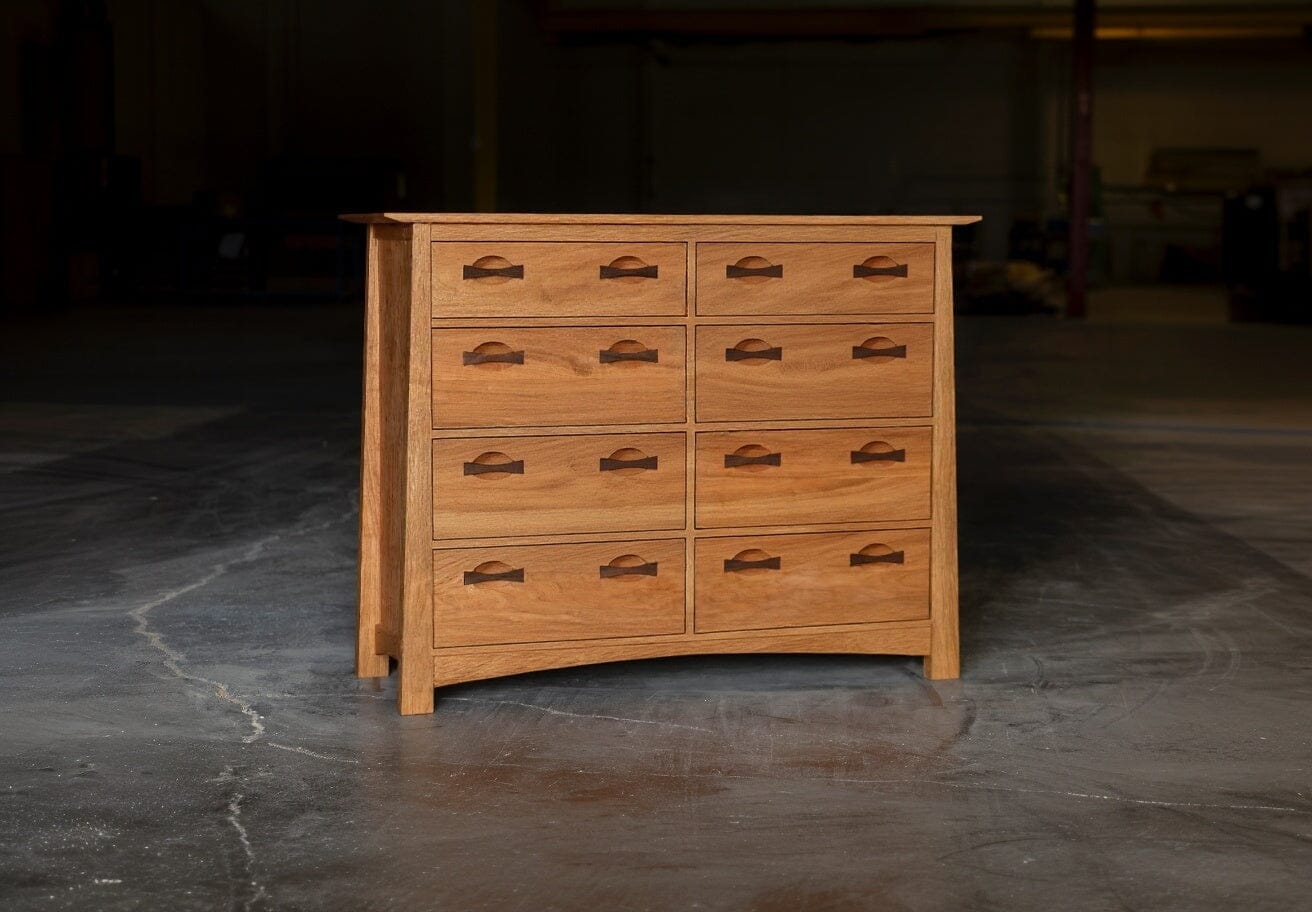 The Enso 6/8 Drawer Dresser by TY Fine Furniture, handmade from solid cherry wood, stands on a concrete floor in a dimly lit room. It features simple handles, a natural finish, and timeless appeal.