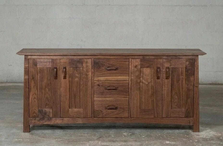 The Enso Credenza & Buffet in Walnut by TY Fine Furniture is a sustainable solid wood piece featuring two side cabinets and three center drawers, shown on a concrete floor against a light-gray wall.