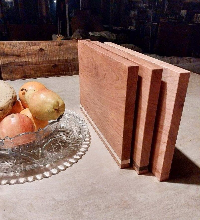 A Handmade Hardwood Cutting Board Set by TY Fine Furniture stands upright on a table beside a glass dish filled with oranges and pears.