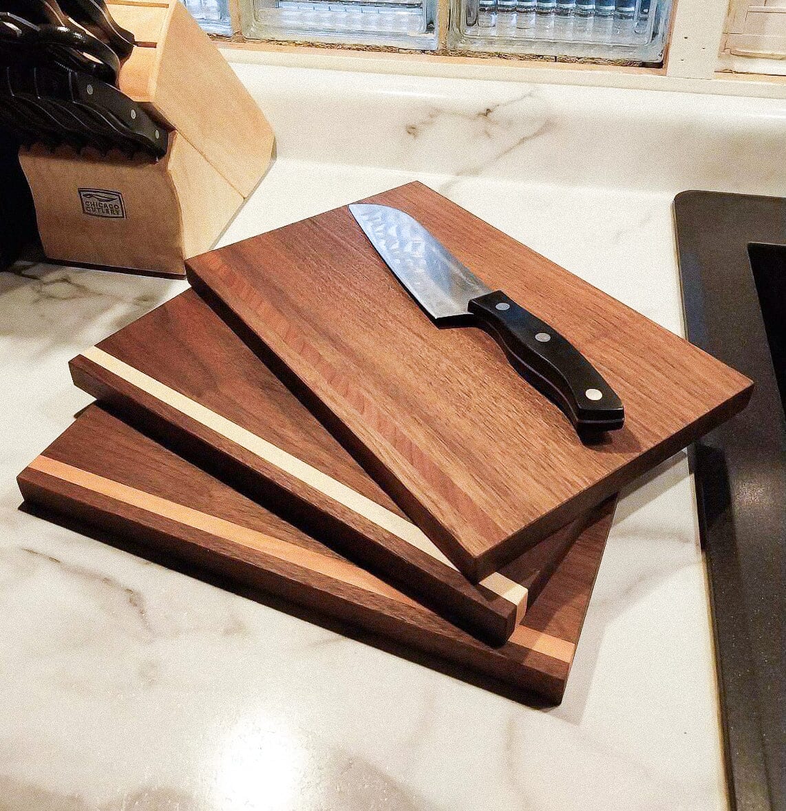 Three Handmade Hardwood Cutting Boards by TY Fine Furniture are stacked on a light marble counter with a chef’s knife on top. A knife block in the background highlights this stylish set.