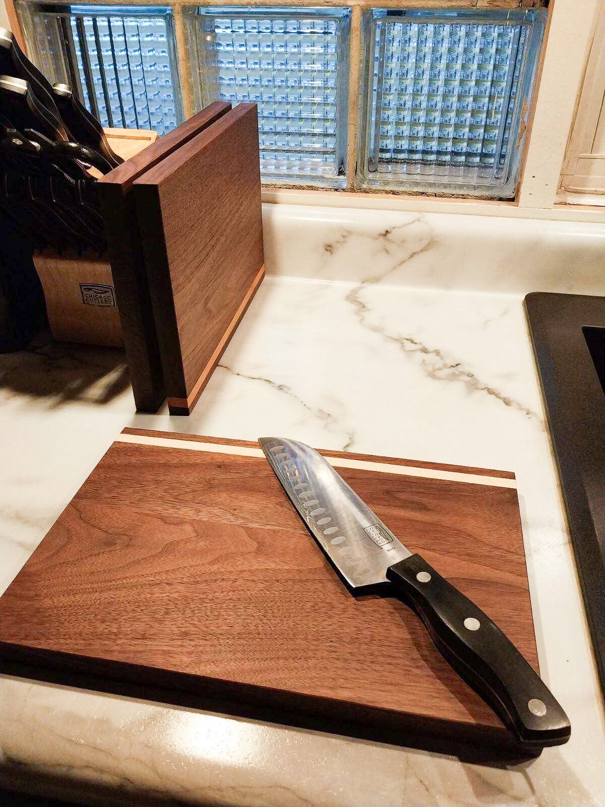 On the kitchen counter sits the TY Fine Furniture Handmade Hardwood Cutting Board Set and a chef's knife; another wooden board and a knife block are near a window with frosted glass blocks in the background.