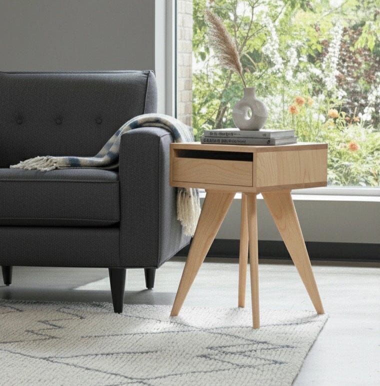 A modern living room features a dark gray sofa, plaid throw, and handcrafted flair with the TY Fine Furniture Modern Apartment Side Table - Maple. The light wood table holds a white vase with dried grass and books. Greenery is visible through large windows.