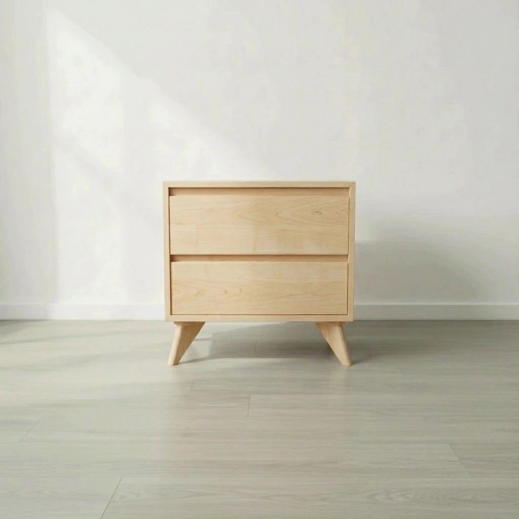 The TY Fine Furniture Modern Cube Side Table - Maple is a small, modern solid wood piece with angled legs and two push-to-open drawers, shown on a light gray wooden floor against a plain white wall.