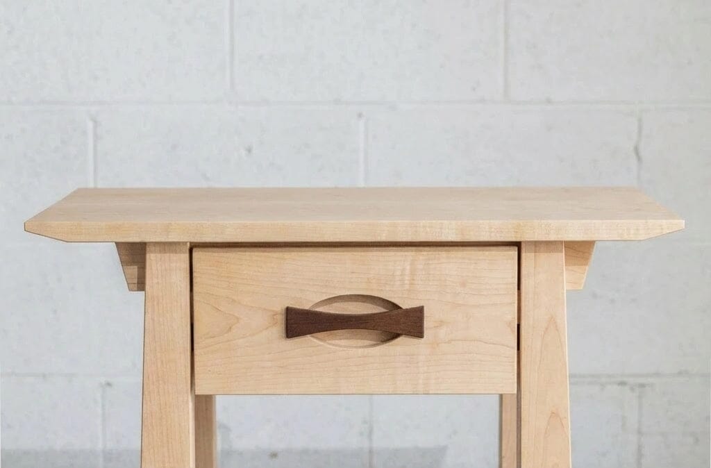 The Enso Side Table with Drawer in Maple by TY Fine Furniture is a handcrafted solid wood piece featuring a unique dark bowtie handle, blending elegance and functionality against a light brick wall.