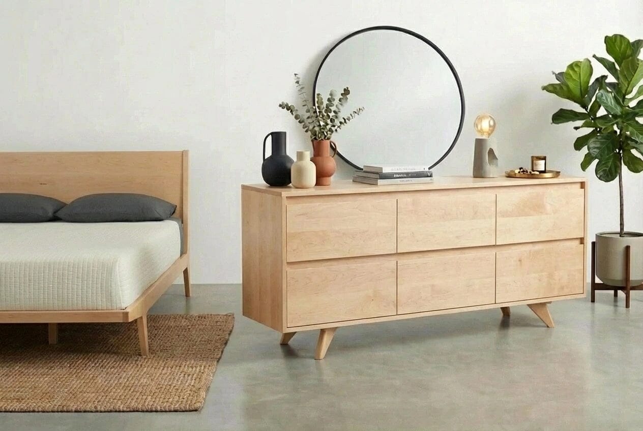 A minimalist bedroom features a wooden bed, TY Fine Furniture’s Modern Mid-Century Dresser in Maple (Columbus), a round mirror, potted plant, books, decorative vases, all set on concrete flooring with a woven rug.