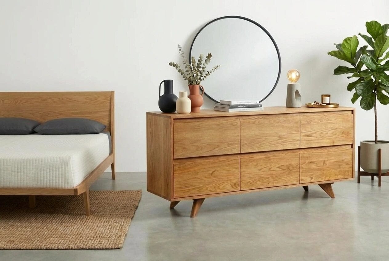 A modern bedroom includes a gray blanket on a wooden bed, TY Fine Furniture’s Modern Mid-Century Dresser in White Oak - Solid Wood | Colum with decor, a round mirror, potted plant, and woven rug on a concrete floor.