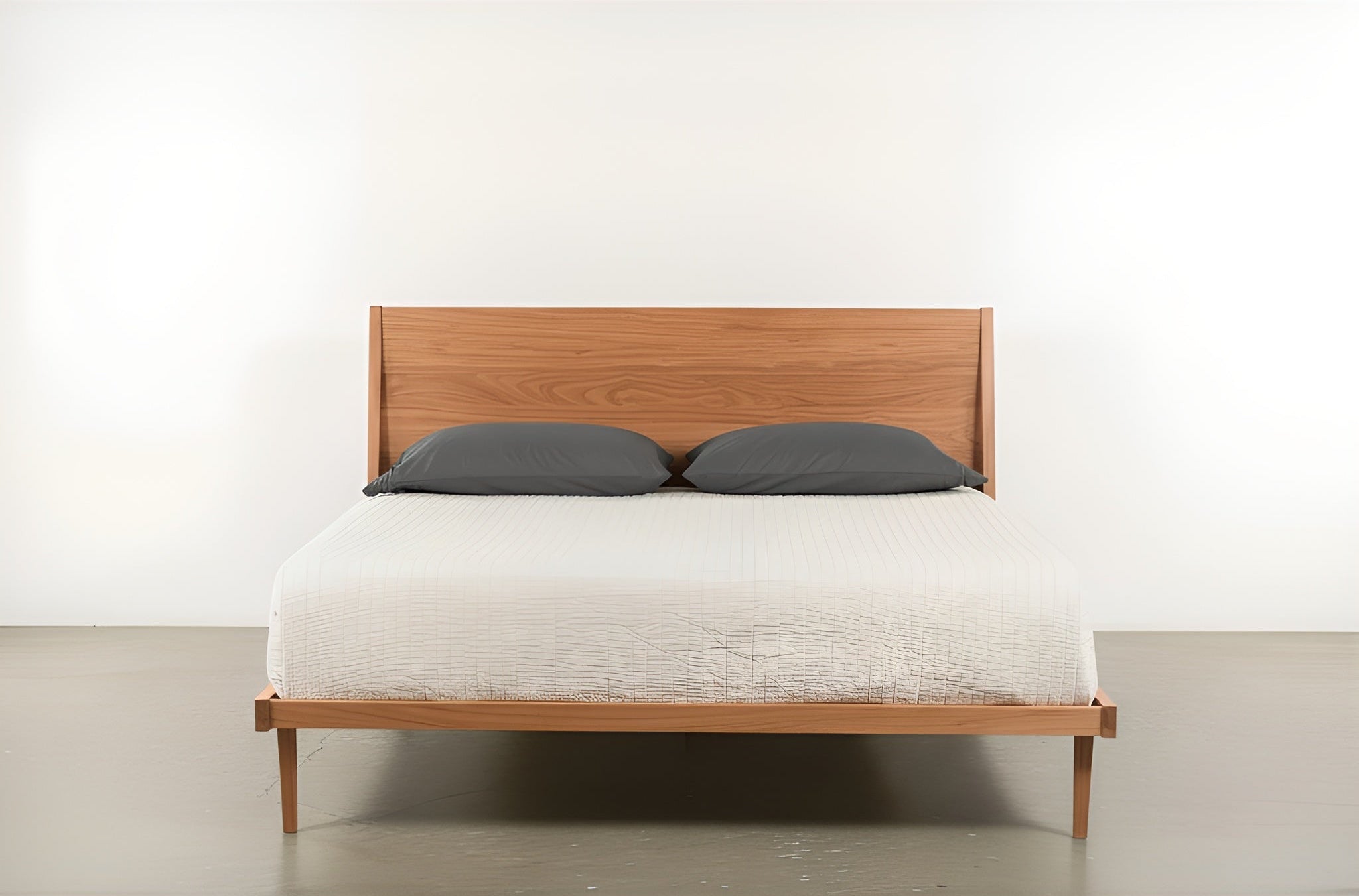 The Modern "ONE" Platform Bed by TY Fine Furniture, featuring a solid wood cherry frame and matching headboard, is topped with a white quilt and gray pillows, and rests against a white wall on a polished concrete floor.