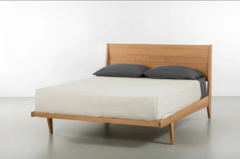 The Modern "ONE" Platform Bed by TY Fine Furniture, handcrafted from solid white oak, features an angled headboard and is styled with a beige quilted bedspread and two dark gray pillows, set against a plain white background.