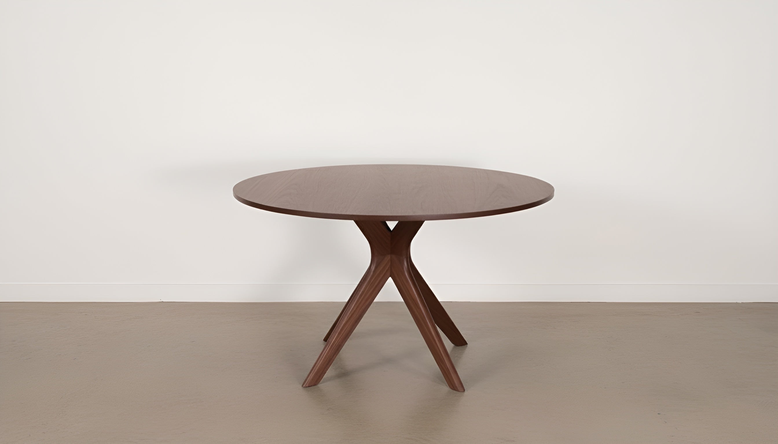 The Strata Round Dining Table by TY Fine Furniture features a smooth, handcrafted solid wood surface and four intersecting legs, shown on a light floor against a plain white wall.