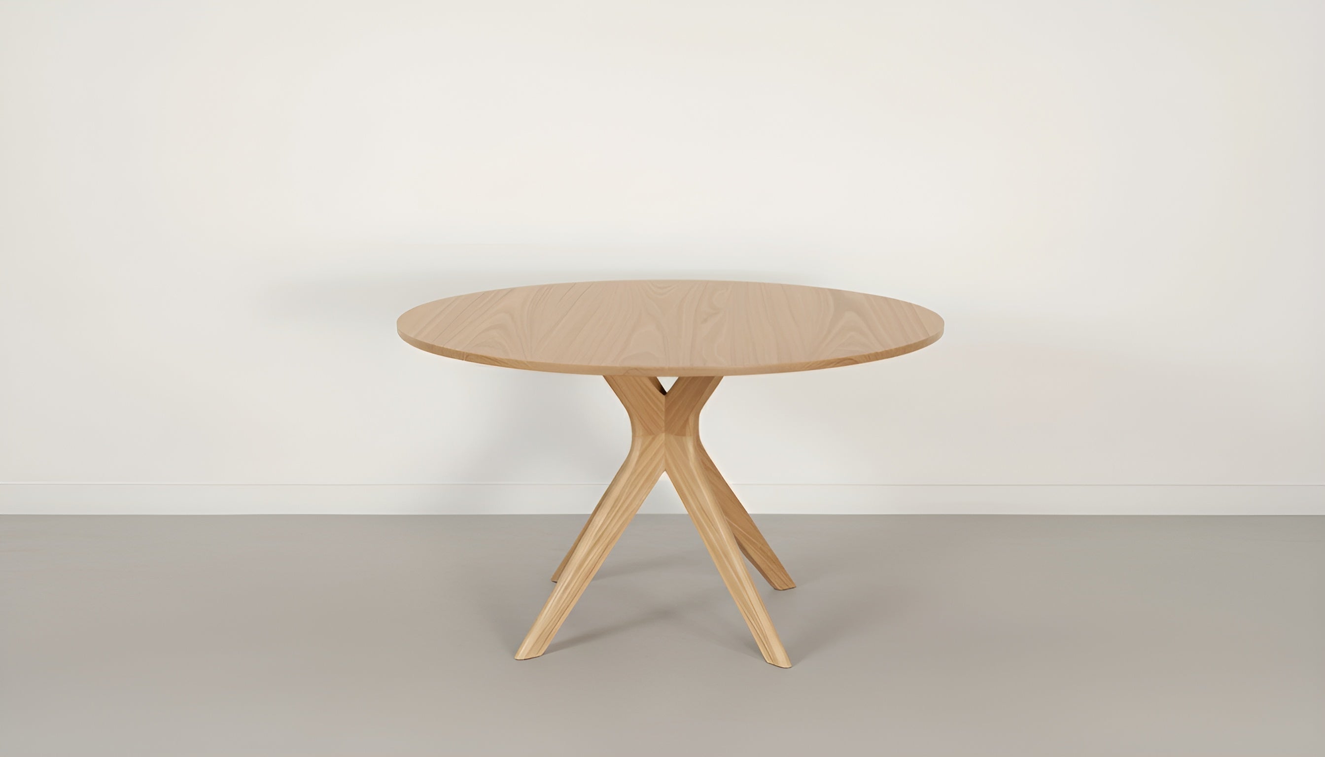 The Strata Round Dining Table by TY Fine Furniture features a smooth, handcrafted solid wood finish and three angled legs, displayed on a light grey floor against a plain white wall.