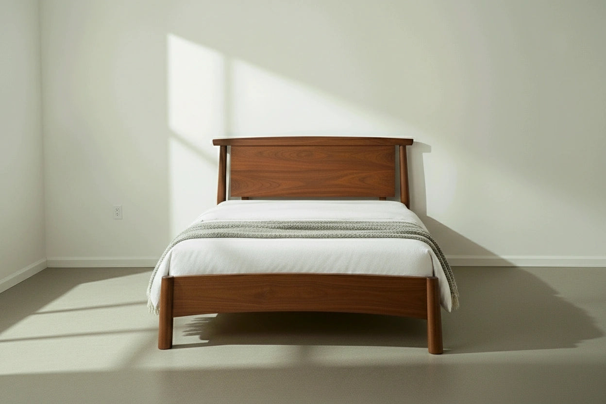 The Sunrise Platform Bed - Walnut by TY Fine Furniture, topped with a white mattress and light gray blanket, stands in a minimalist room with white walls and sunlight streaming in, creating gentle shadows on the floor.