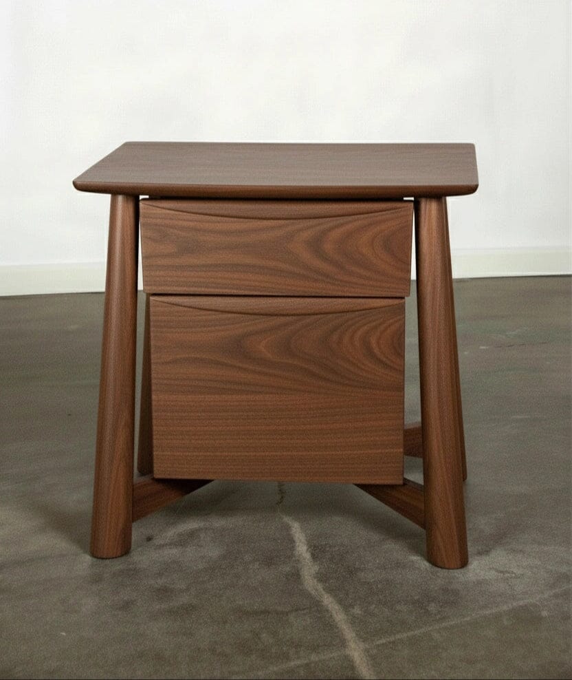 The Sunrise Side Table - Walnut by TY Fine Furniture, a handcrafted modern piece with angled legs, two drawers, smooth finish, and visible wood grain, sits on a concrete floor against a plain light-colored wall.
