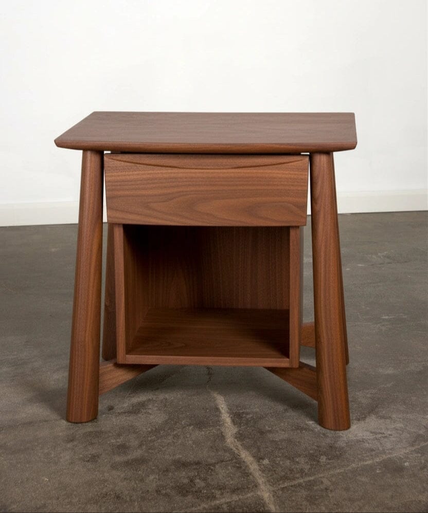 The Sunrise Side Table - Walnut by TY Fine Furniture, made from natural solid wood with angled legs, a small drawer, and an open lower shelf, stands on a concrete floor against a plain white wall.