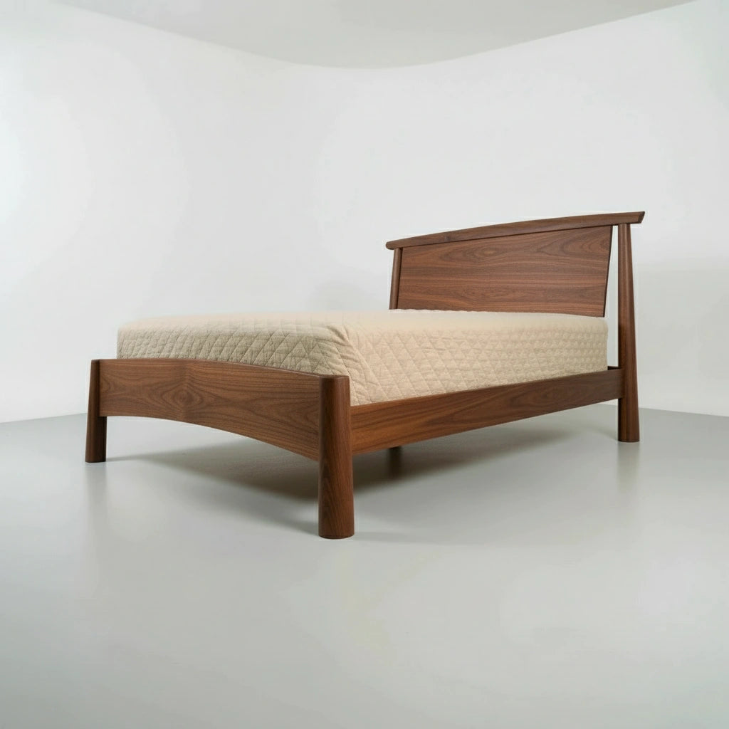 The Sunrise Platform Bed - Walnut by TY Fine Furniture features a handcrafted solid wood frame, angled legs, and a subtly curved headboard, complemented by a beige quilted mattress in a minimalist, white-walled room.