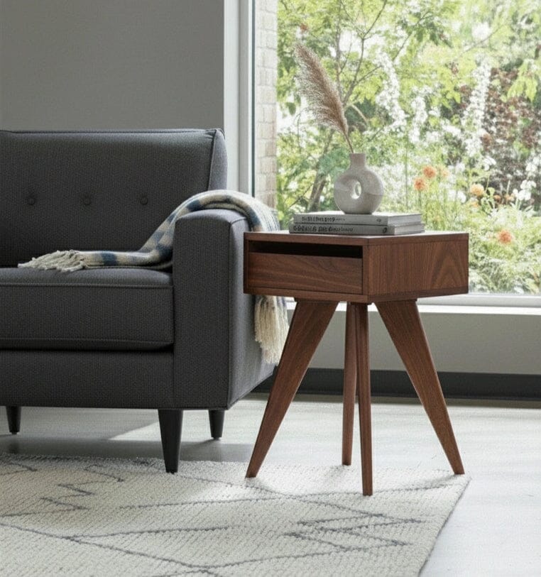 A modern living room features a dark gray sofa with a plaid blanket, and the TY Fine Furniture Modern Apartment Side Table in walnut displaying a vase of dried grass and two books. A large window showcases lush greenery outside.
