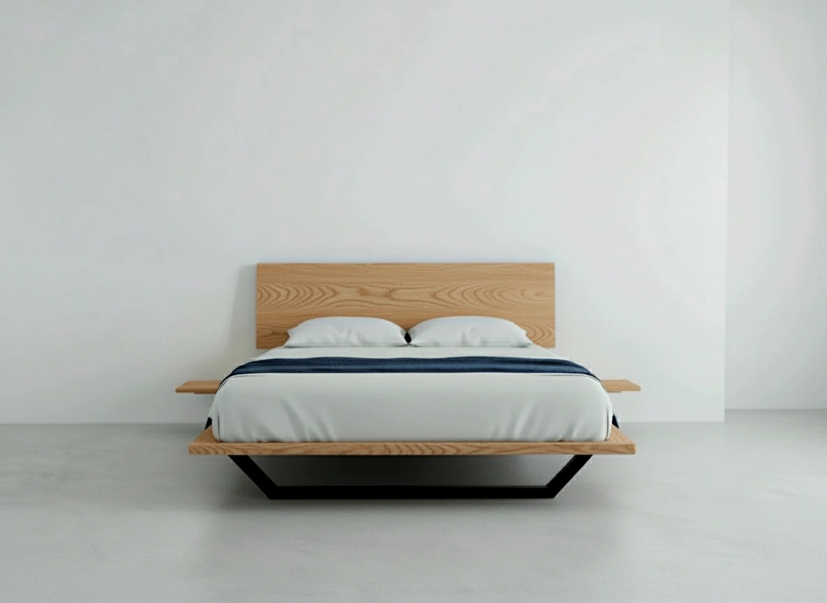 The Nelson Solid Wood Platform Bed Frame by TY Fine Furniture, crafted from American white oak, features a low oak frame, solid oak headboard, white mattress and bedding, two pillows, and a dark blue blanket against a plain white wall and floor.