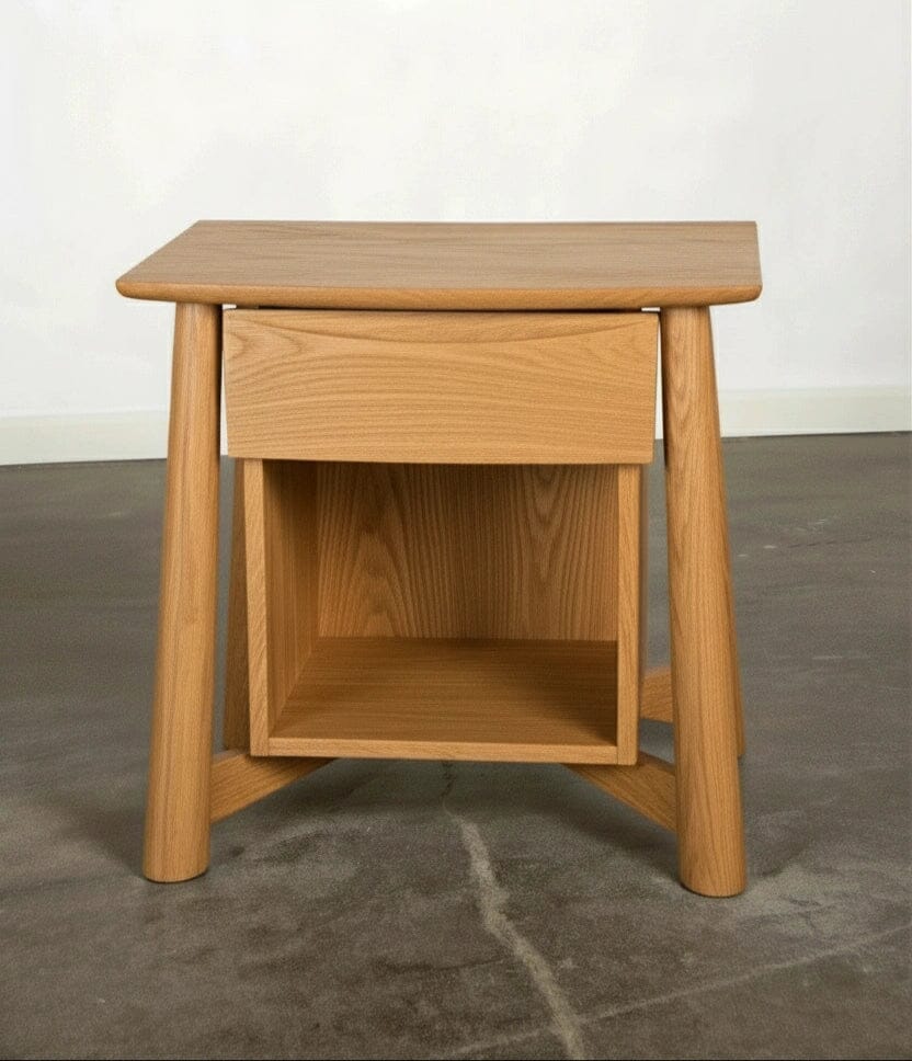 The Sunrise Side Table - White Oak by TY Fine Furniture is a handcrafted modern piece with angled legs, a single drawer, and an open lower shelf, made of solid wood and shown on a concrete floor against a plain white wall.