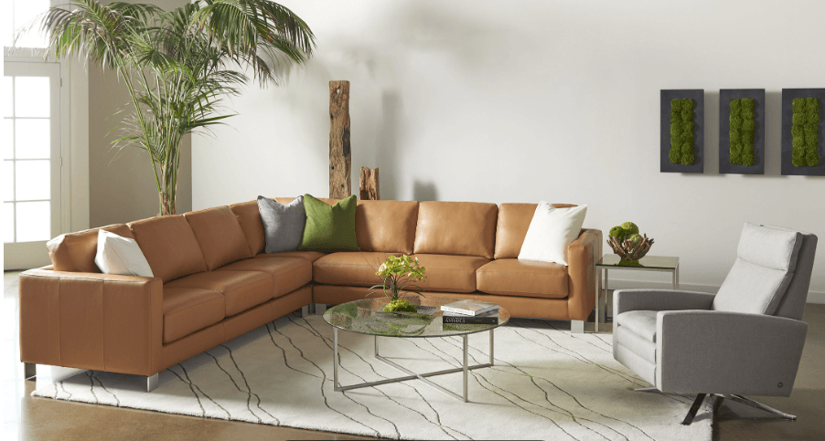 A modern living room showcases the American Leather Alessandro Sofa, contemporary loveseat and chair, all with high-resiliency foam cushions, plus a gray armchair, glass coffee table, potted plant, green wall art, and a geometric area rug.