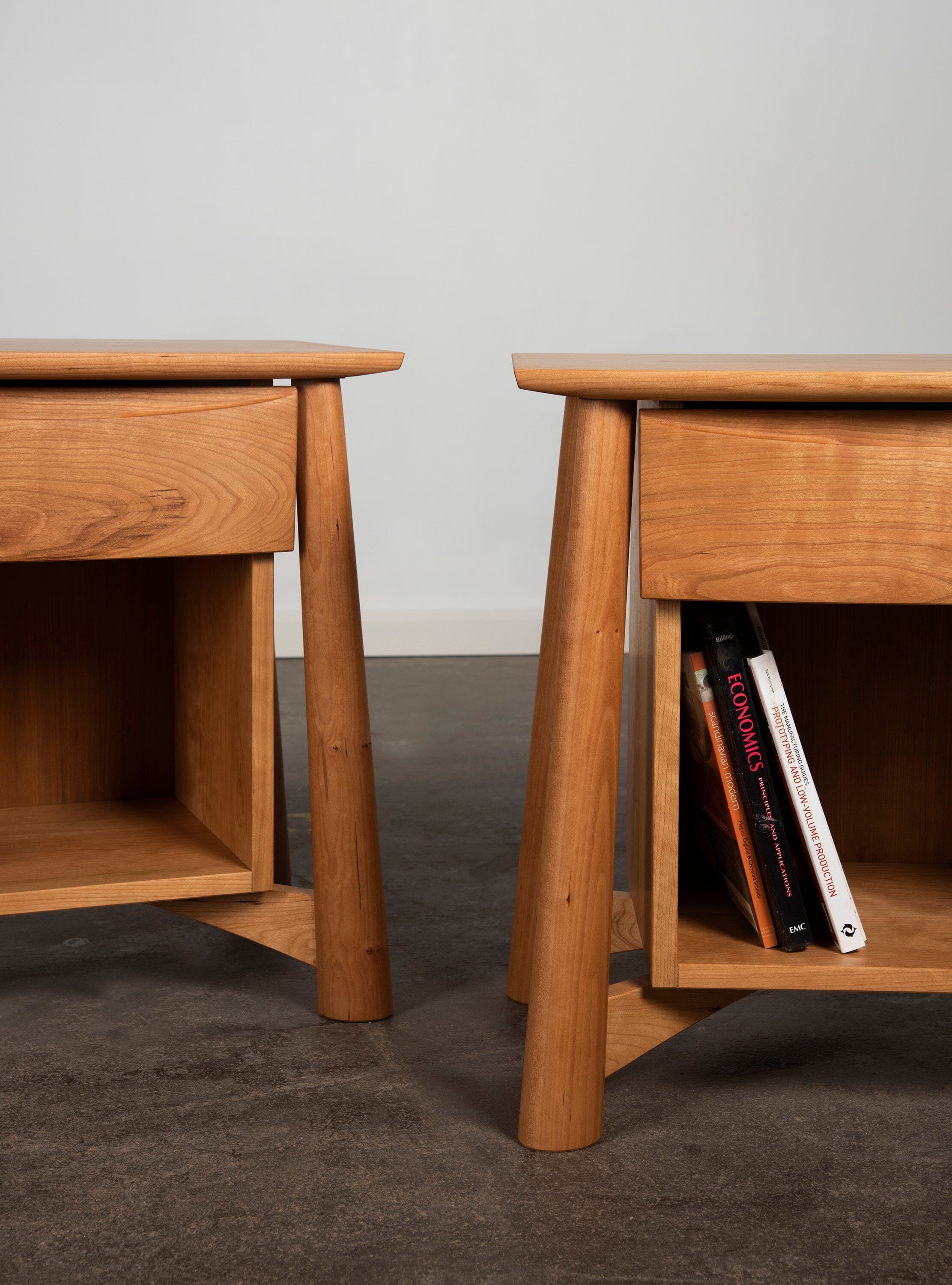Two TY Fine Furniture Sunrise Side Tables in cherry, both with light natural finishes and angled legs, stand on a dark floor. One holds books on its lower shelf while the other is empty.