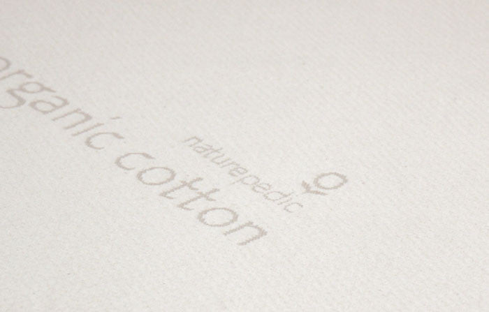 Close-up of a white fabric surface with faint printed text reading "organic cotton" and "Naturepedic," plus a small logo—highlighting the quality of the Naturepedic Organic Latex Mattress Topper, designed for pressure point relief.