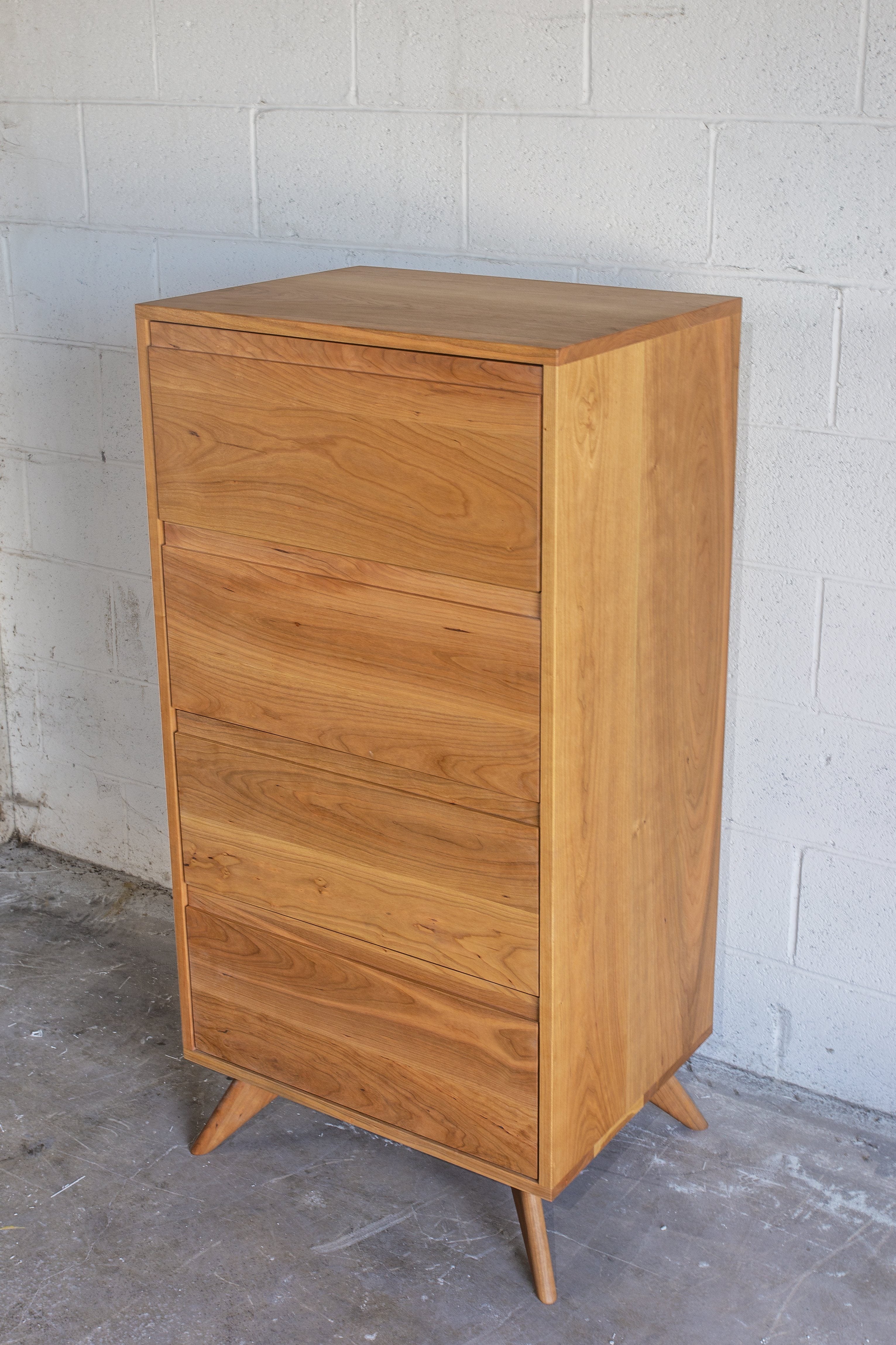 The TY Fine Furniture Tall Modern Dresser Mid Century- Cherry features five wooden drawers and angled legs, standing stylishly against a light gray concrete block wall on a cement floor.