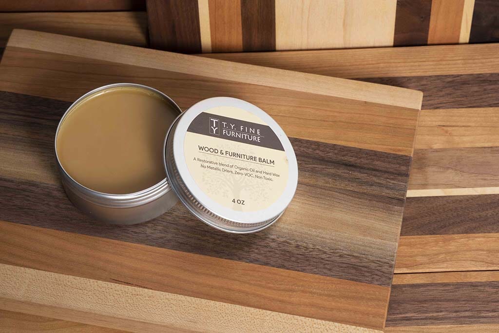 An open tin of TY Fine Furniture Wood and Furniture Balm—Organic Hard Wax Wood Finish—rests on a striped wood surface. The smooth, light brown balm restores and protects your furniture’s finish.
