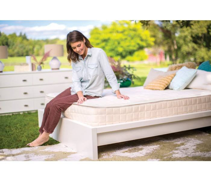 A woman sits outdoors on a Naturepedic Chorus Mattress with white bedding and pillows, smiling and touching the organic mattress. The bed is surrounded by greenery and trees, with a white dresser visible nearby. Brand: Naturepedic.