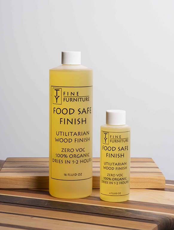 Two bottles of TY Fine Furniture’s Wood Finish Organic All Natural - Oil Blend (16 oz and 4 oz) sit on a wooden cutting board. Both highlight zero VOC, 100% organic ingredients, and quick drying for easy furniture refinishing.