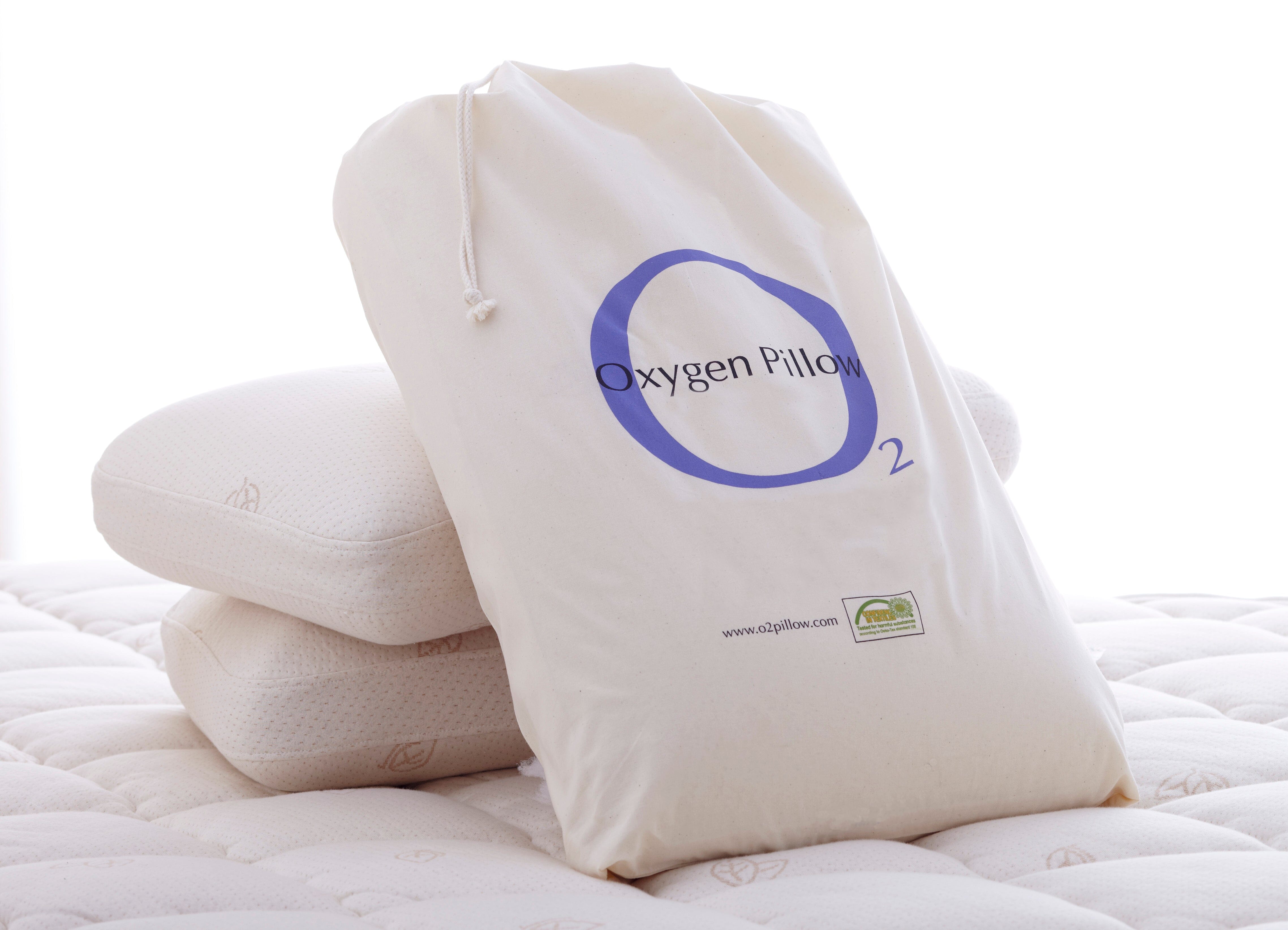 A white drawstring bag labeled Oxygen Pillow - Natural Fill | TY Sleep by TY Sleep sits upright on a mattress beside two stacked, subtle-patterned, white pillows—ideal for side sleepers who need edge support.