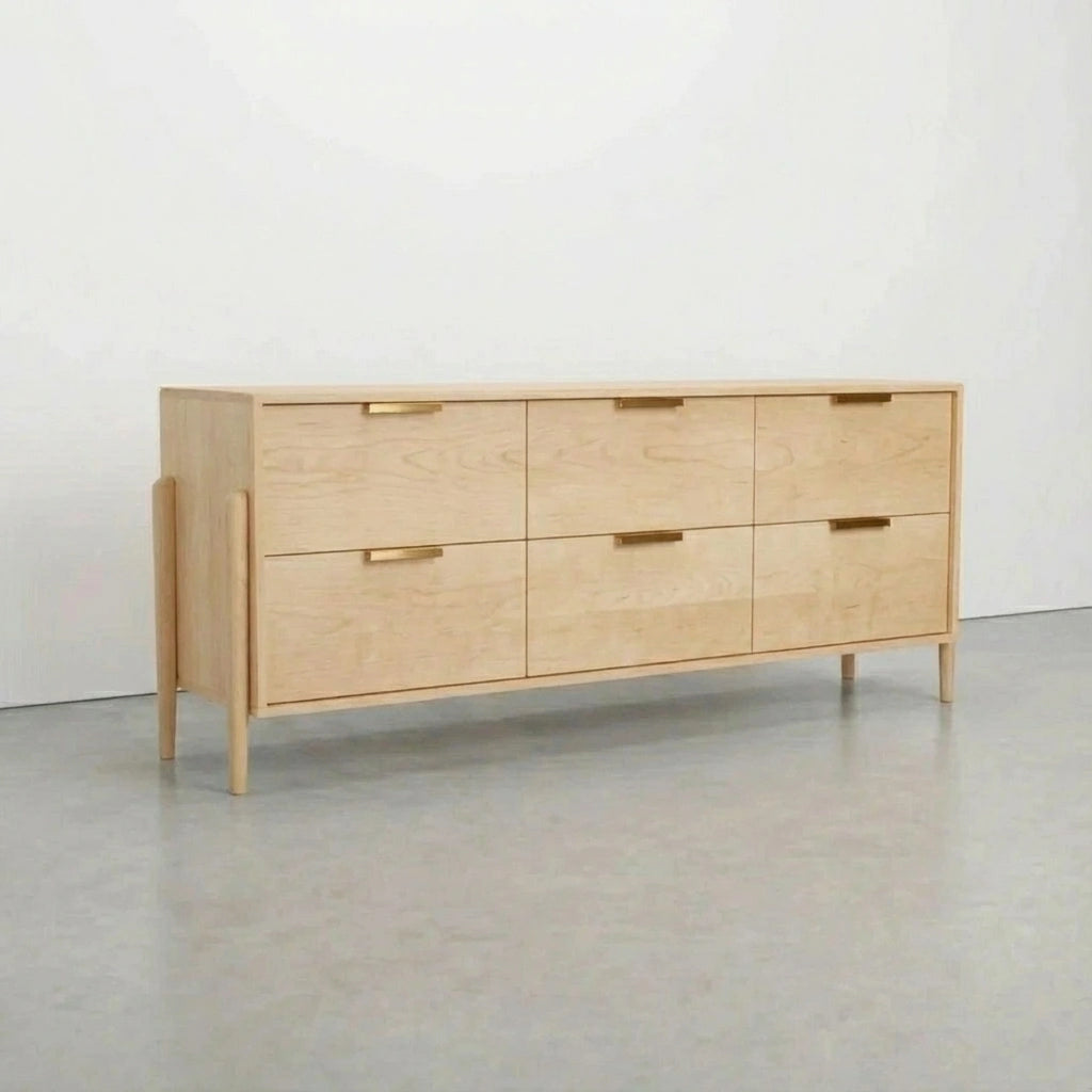 The TY Fine Furniture Apollo 6 Drawer Dresser, a handcrafted maple piece in mid-century modern style, with six drawers and minimalist gold handles, stands on four tapered legs against a white wall and gray floor.