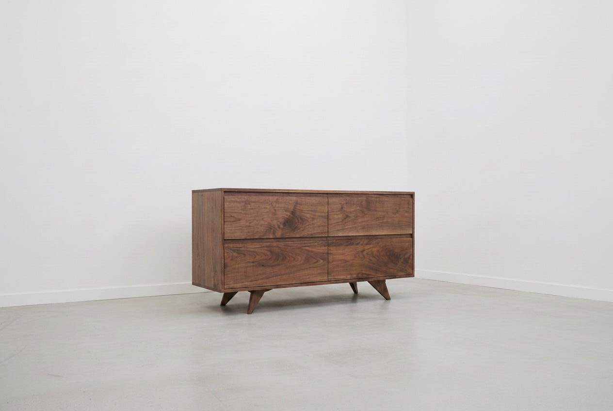 The TY Fine Furniture Modern Mid-Century Dresser in Walnut, a solid wood piece with four drawers and angled legs, stands in an empty room with white walls and a light gray floor.