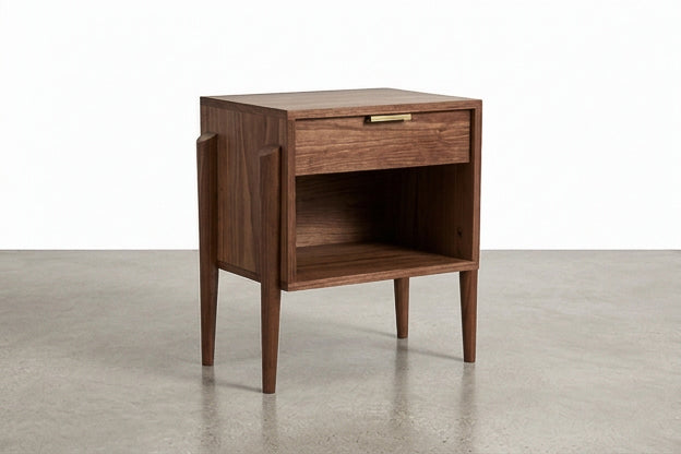 Introducing the Apollo Cube Side Table in Walnut by TY Fine Furniture—a solid hardwood, handcrafted piece featuring a mid-century modern design with tapered legs, a single drawer adorned with a brass handle, and an open shelf below. Set on a smooth concrete floor against a plain white background, this versatile nightstand from Columbus, OH adds timeless style to any interior.