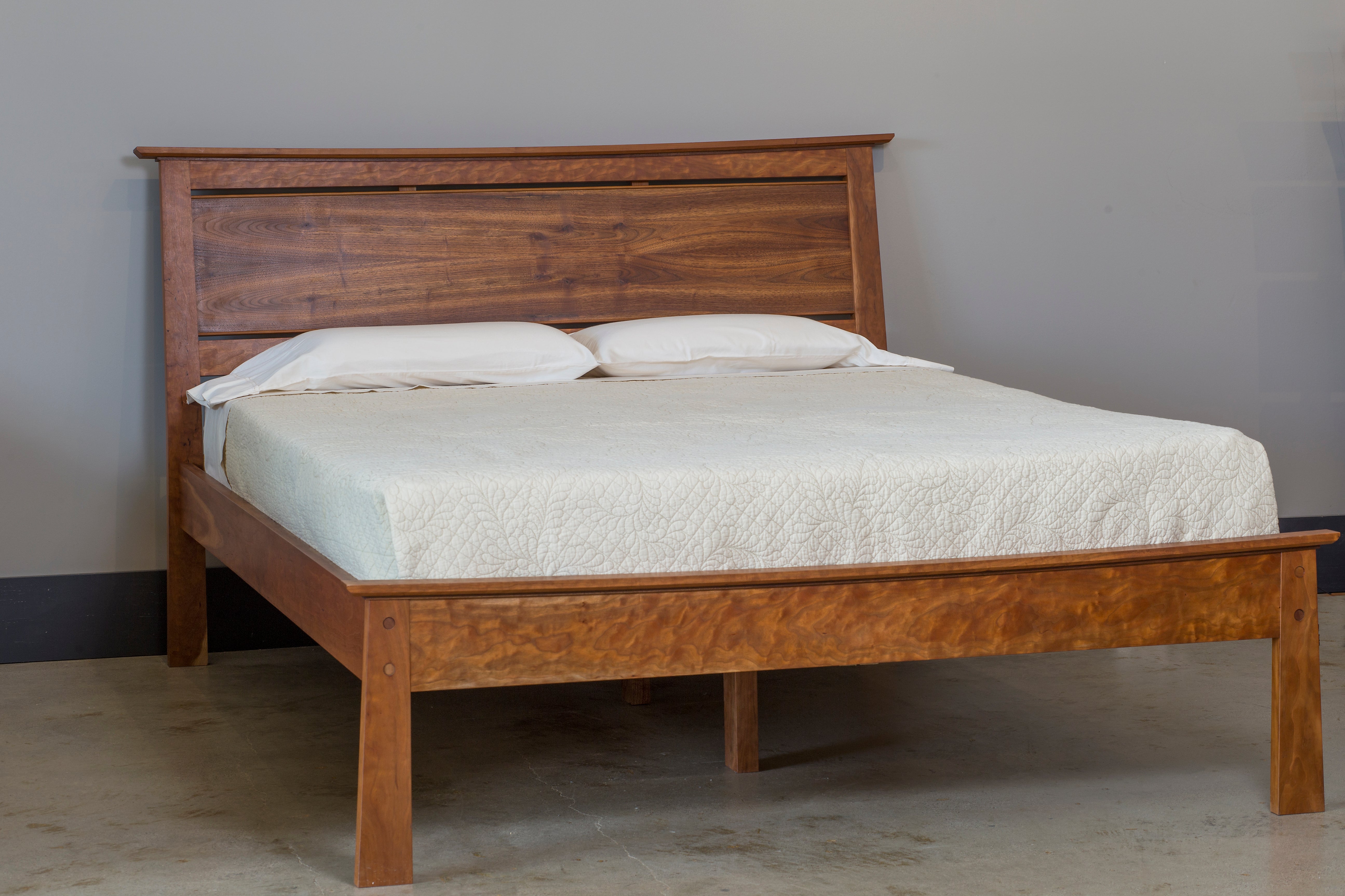 The Enso Platform Bed Walnut by TY Fine Furniture, featuring a high paneled headboard and dressed in a light quilt with two white pillows, rests on a concrete floor against a plain gray wall.