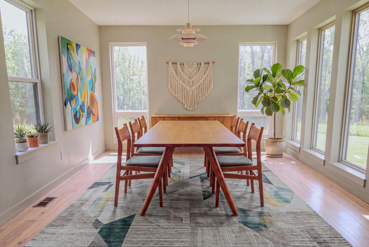 A bright dining room featuring the Solid Cherry Dining Table from the Essentials Collection by TY Fine Furniture with eight chairs, large windows, a potted plant, abstract wall art, and a macrame wall hanging above the table. Light wood floors and a patterned rug complete the decor.
