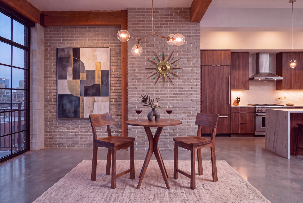 A modern dining area featuring two wooden chairs and the Solid Cherry Round Pub Table from the Strata Collection by TY Fine Furniture, set with wine glasses in a loft with brick walls, abstract art, a sunburst clock, and a kitchen visible in the background.
