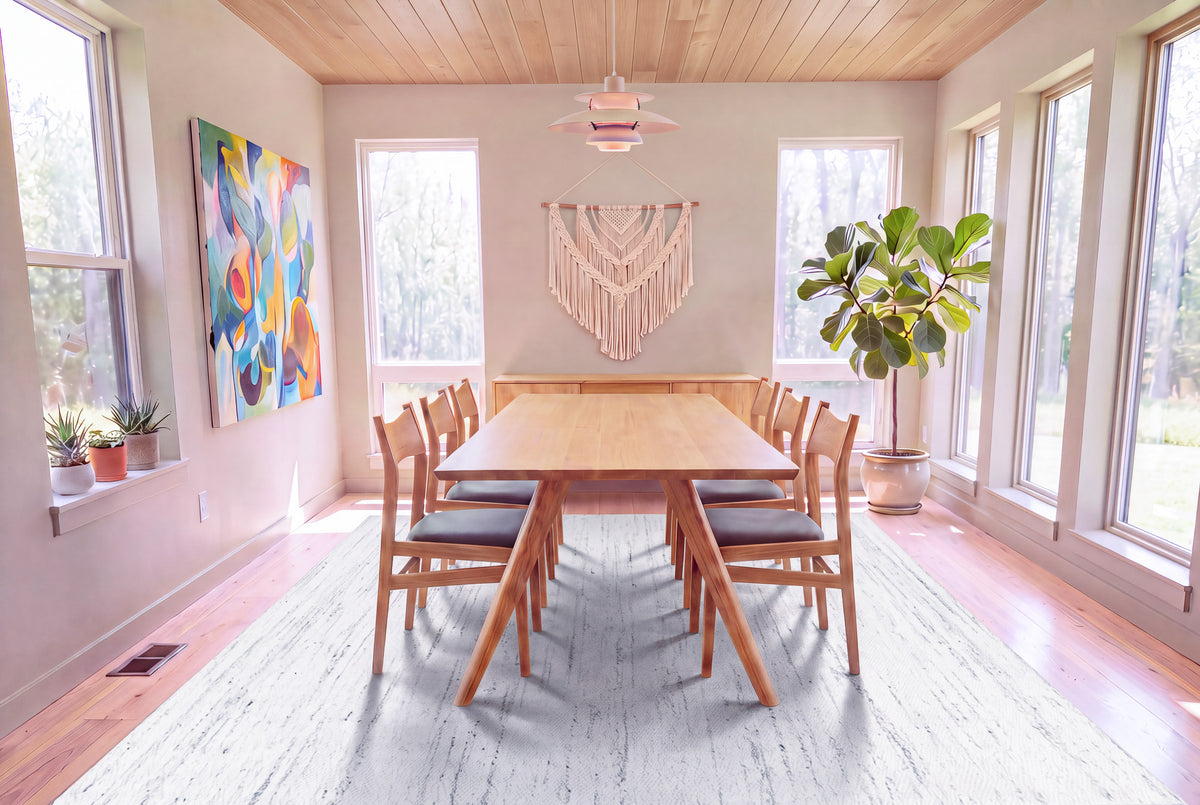A modern dining room highlights the Solid White Oak Dining Table from the Essentials Collection by TY Fine Furniture, surrounded by eight chairs and accented with abstract art, a macramé wall hanging, potted plants, and large windows that fill the space with natural light.