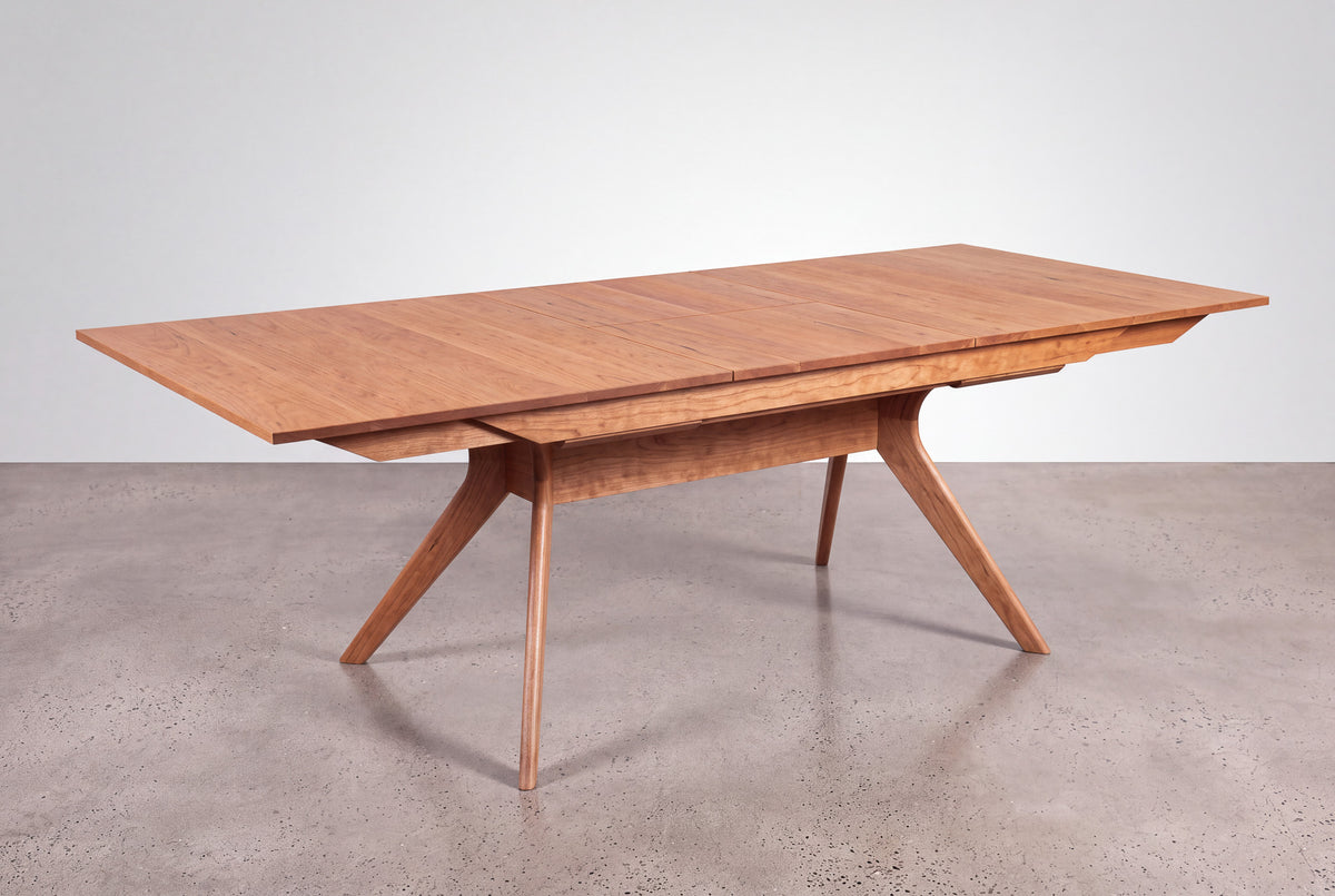 The Solid Cherry Extendable Dining Table from the Strata Collection by TY Fine Furniture, featuring an extended leaf, angled legs, and a natural finish, stands on a polished concrete floor against a plain white background.