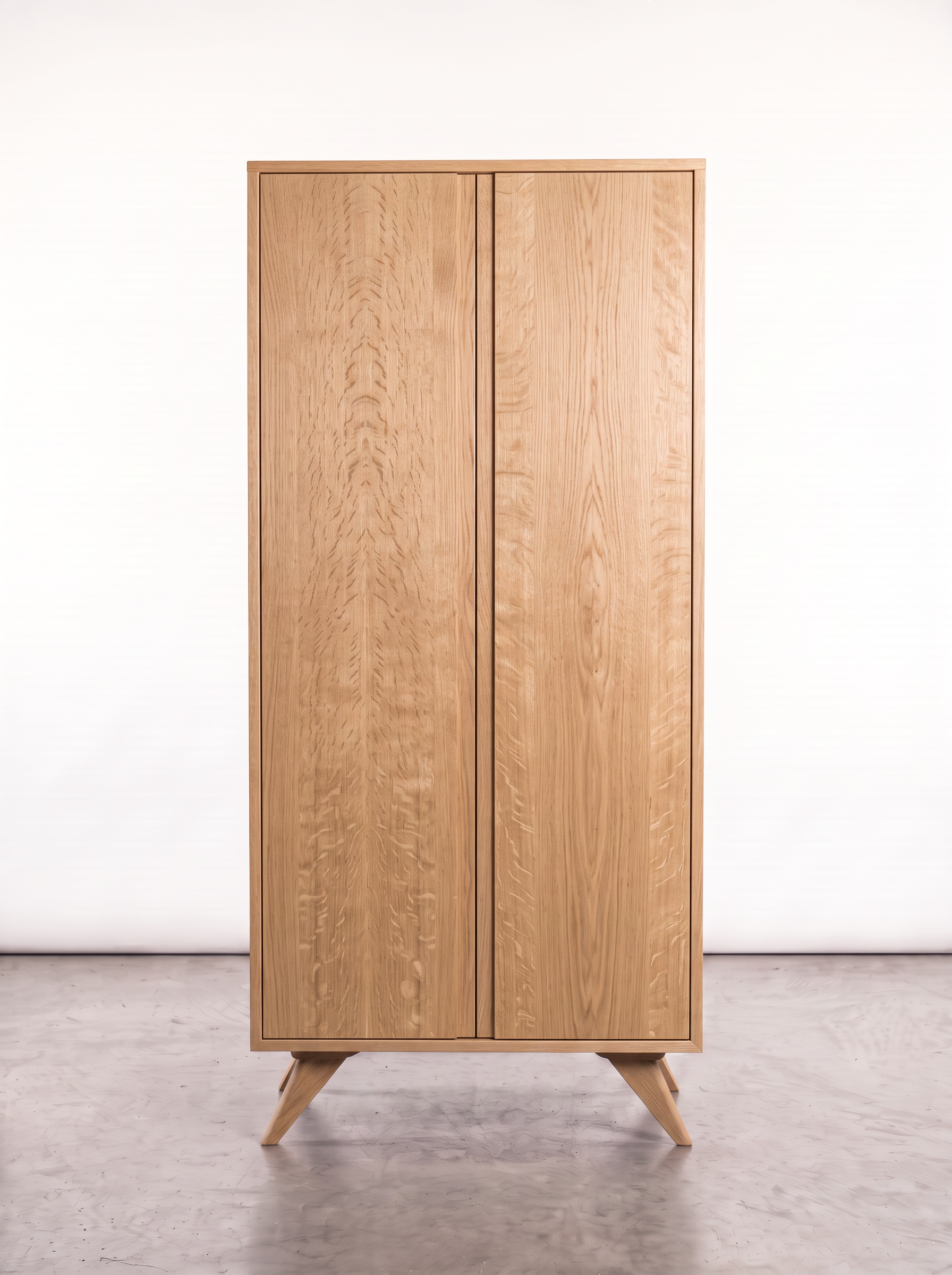The Solid White Oak Tall Storage Cabinet from the Modern Collection by T.Y. Fine Furniture, featuring two doors and four angled legs, is shown standing on a concrete floor against a plain white background.