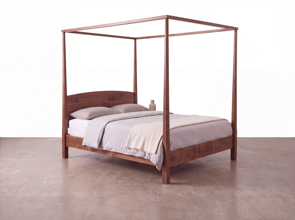 A minimalist Solid Walnut Pencil Post Canopy Bed from the Shaker Collection by TY Fine Furniture, featuring a solid walnut wood frame, is dressed in neutral linen bedding with two pillows and a small ceramic vase, set against a white background on a concrete floor.