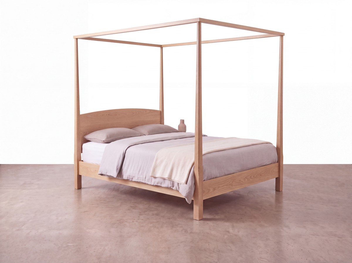 A Solid White Oak Pencil Post Canopy Bed from the Shaker Collection by TY Fine Furniture features light gray bedding and a beige blanket. A small vase is placed on the bed near the headboard, set against a minimal, bright background with a smooth, light brown floor.