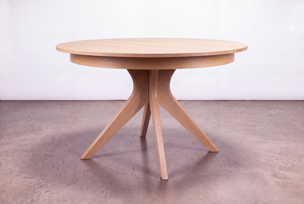 Introducing the Solid White Oak Extendable Round Dining Table from TY Fine Furniture’s Strata Collection—this handcrafted piece features four angled legs and stands elegantly on a polished concrete floor against a simple light background.