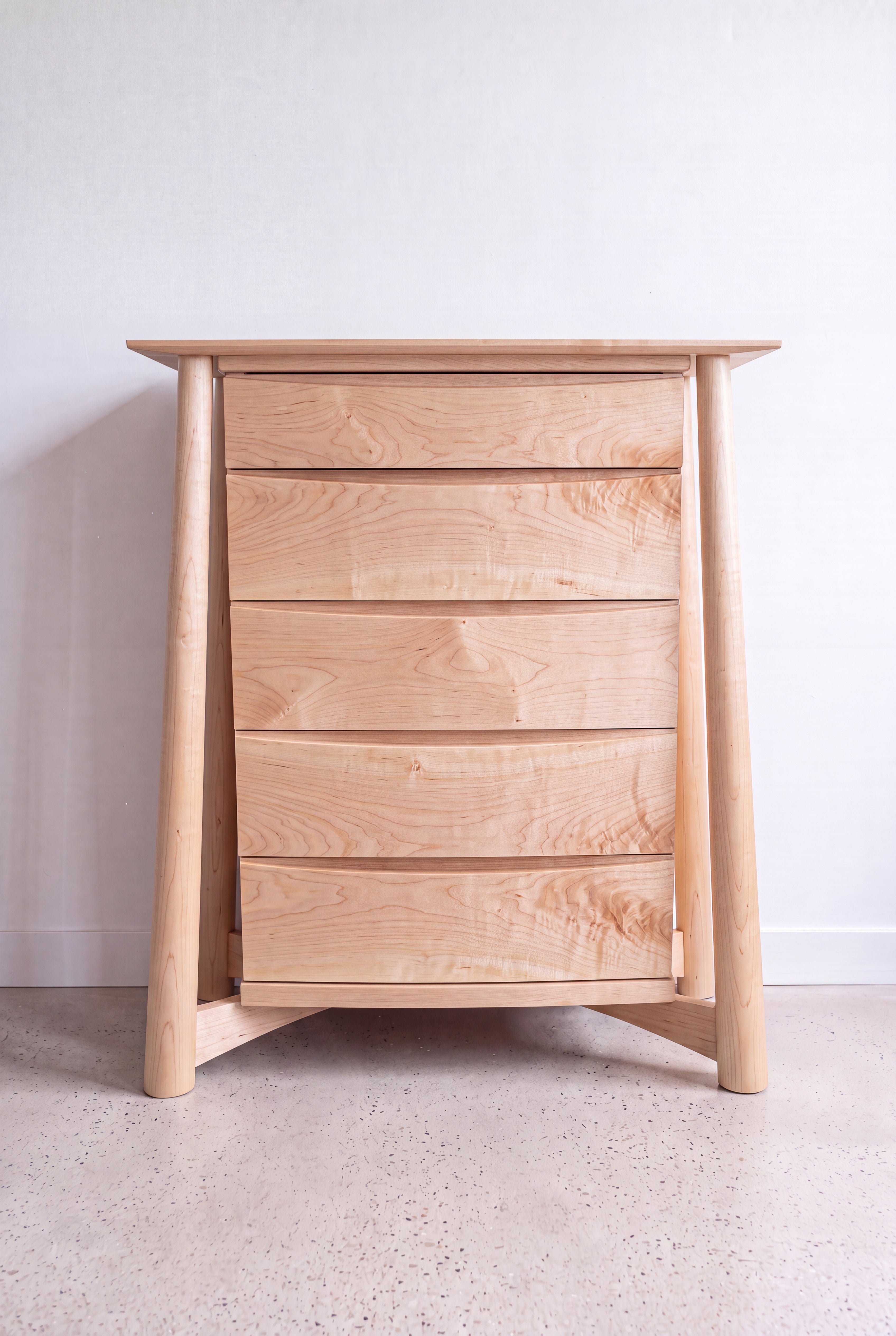 The Sunrise High Dresser by TY Fine Furniture, a handcrafted solid wood white oak dresser with four drawers, angled side legs, and a modern minimalist design, stands against a pale wall on a speckled floor.
